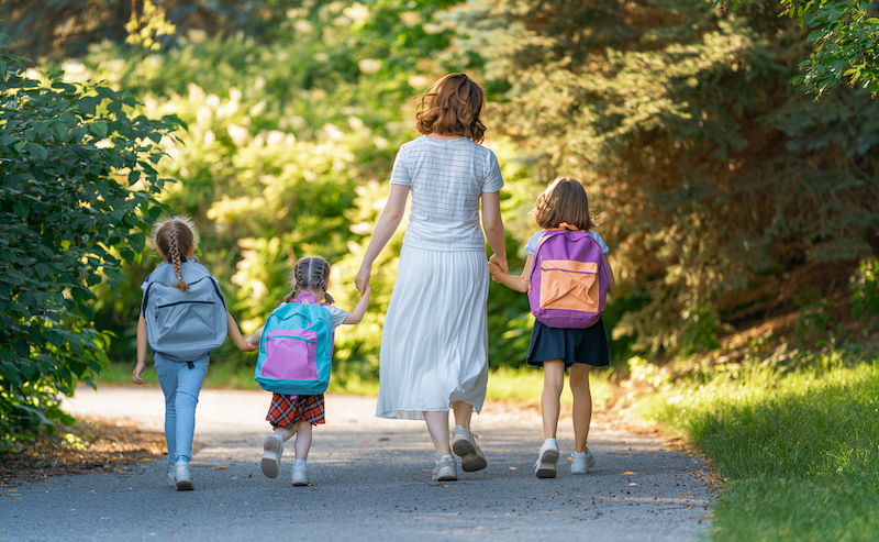 Parent and pupils of primary school go hand in hand. Woman and girls with backpacks behind backs. Beginning of lessons. First day of fall.