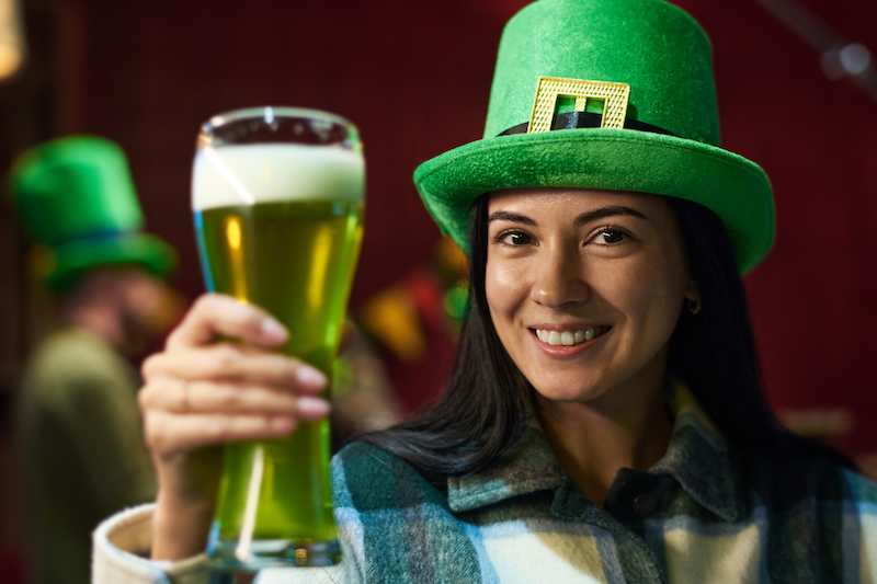 Happy young brunette woman in green hat looking at camera with toothy smile and toasting with glass of foaming beer at party in Irish pub