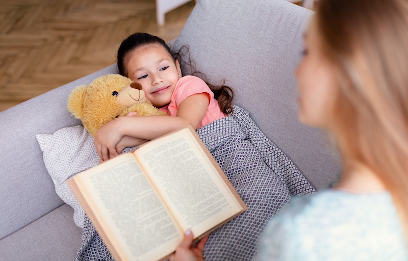 Bedtime Story. Mother Reading Book For Little Daughter While Kid Lying On Couch Hugging Teddy Bear At Home. Selective Focus