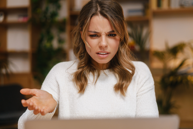 Frustrated confused young woman manager looking at laptop computer while sitting at the office desk, gesturing