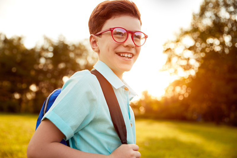 Optimistic little nerd in glasses smiling and looking away while spending time in park after studies at sunset