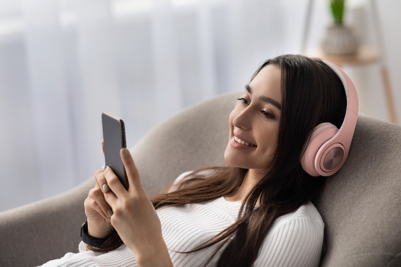 Online lesson remotely, enjoy listening to music, learn language with devices. Glad arab female in headphones typing message on smartphone, watch funny video in armchair in living room interior