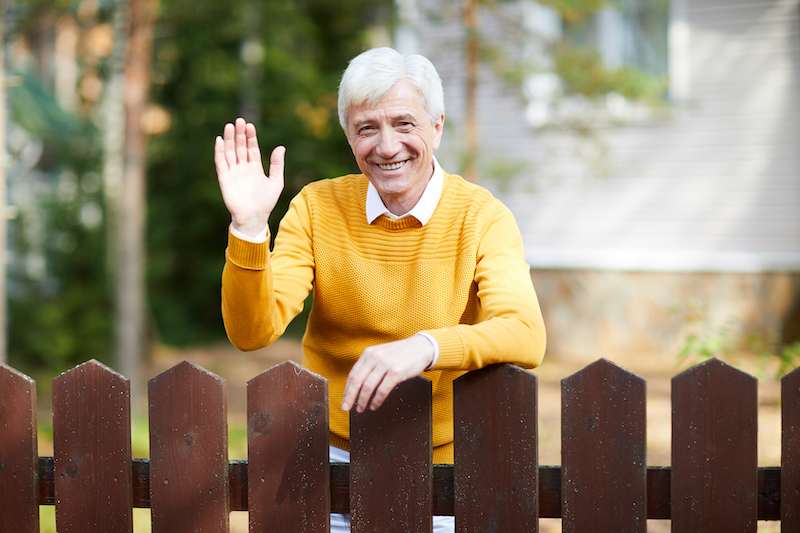 Cheerful senior man in casualwear standing by wooden fence and waving his hand to you