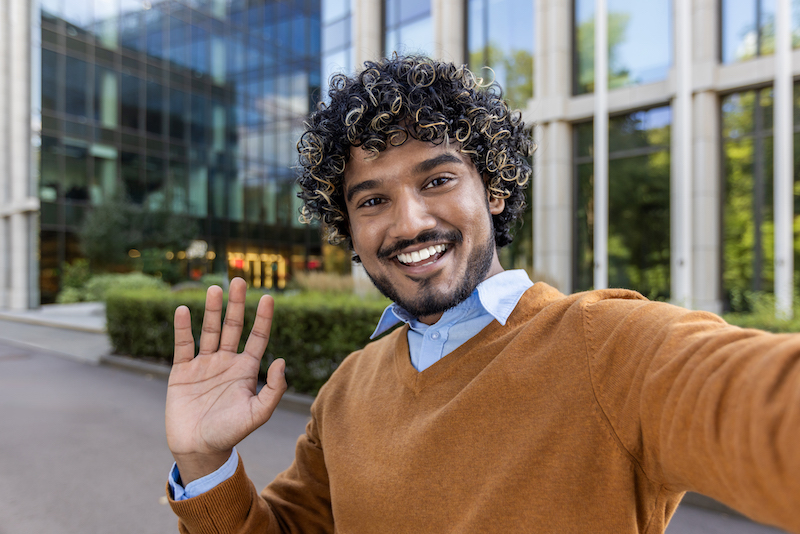 Cheerful man taking selfie outside modern office building. Man is smiling, waving, creating welcoming atmosphere. Bright sunny day enhances positive and professional mood.