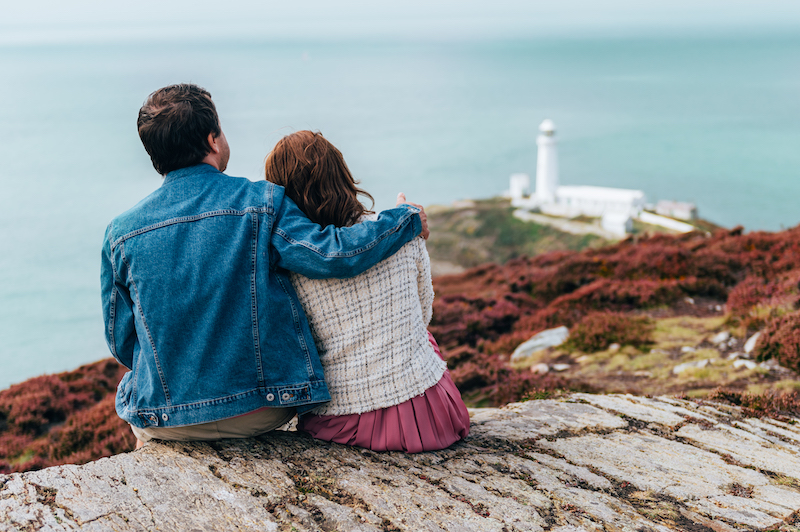 Couple is sitting on the edge of cliff, South Stack Lighthouse on background, Wales, Anglesey. Staycation and travelling around UK.