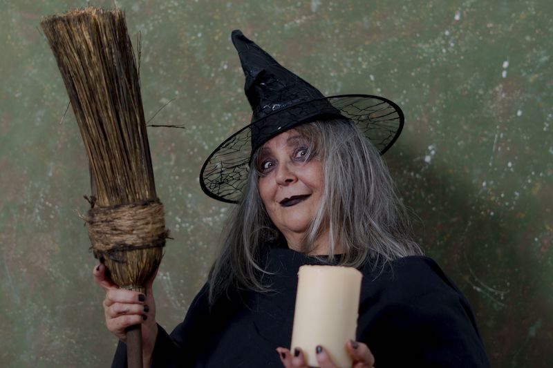 A closeup of a white-haired witch in a black hat and costume holding a broom