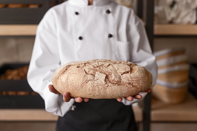 Kid in chef's uniform demonstrating loaf of fresh bread while standing near shelves in bakery