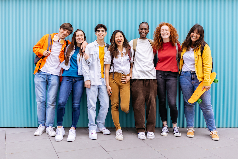 Portrait of seven young student friends leaning on blue wall.