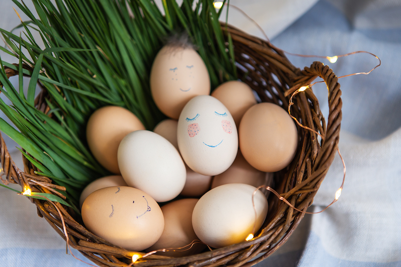 Beautiful painted Easter eggs with a cute face that lie in a wooden basket decorated with warm lights.