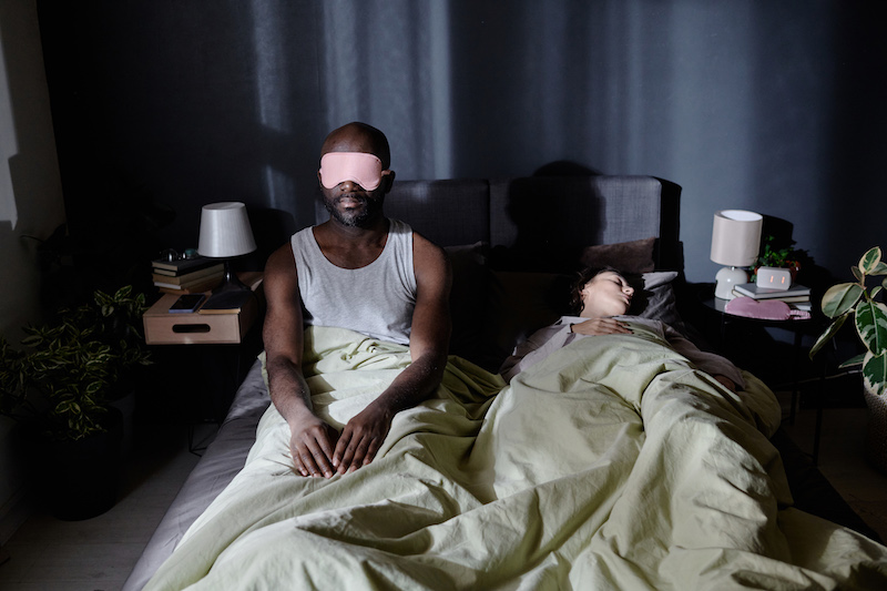 African American man with sleep mask getting up at night while his wife sleeping easy, he suffering from sleepwalking