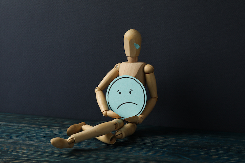 Wooden man with sad emoji in hands on dark blue background