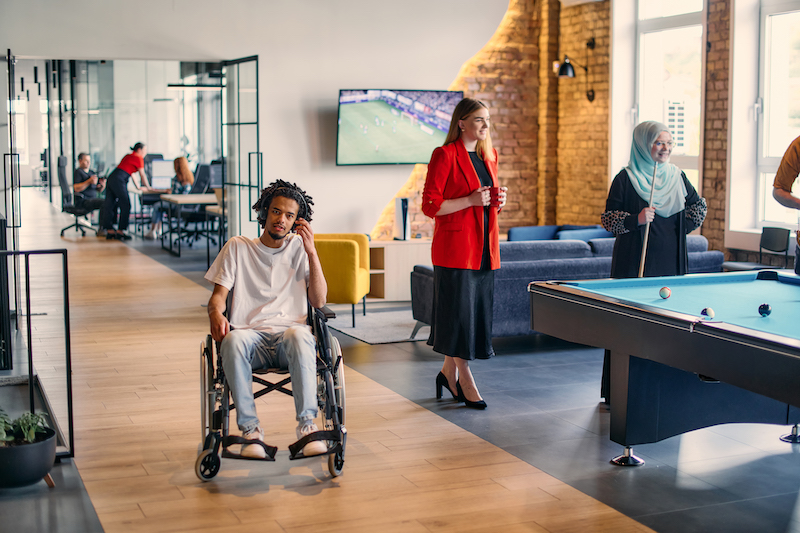 An African-American young entrepreneur in a wheelchair is surrounded by his business colleagues in a modern office setting, embodying diversity and collaboration in the workplace.