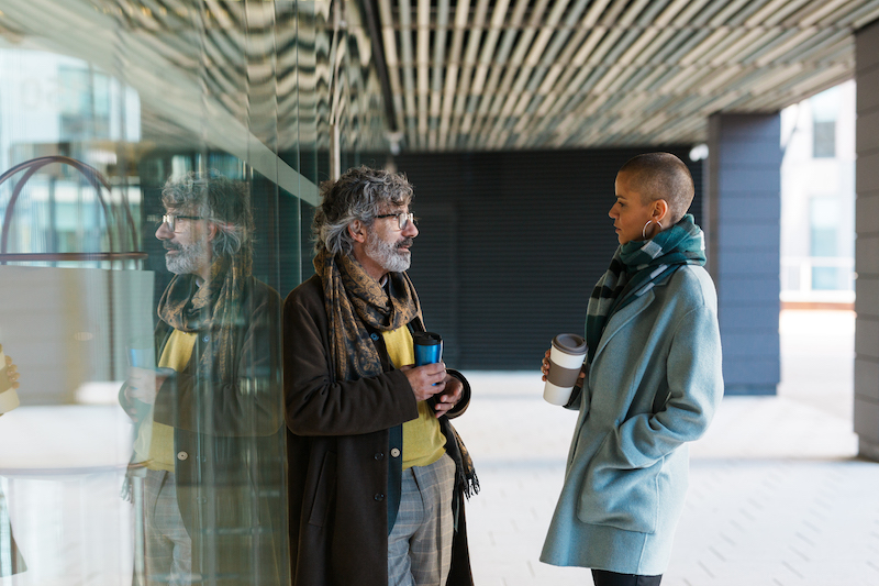 Hispanic man and woman in stylish outerwear enjoying takeaway coffee and talking with each other, while standing near glass wall of modern office building in morning before work
