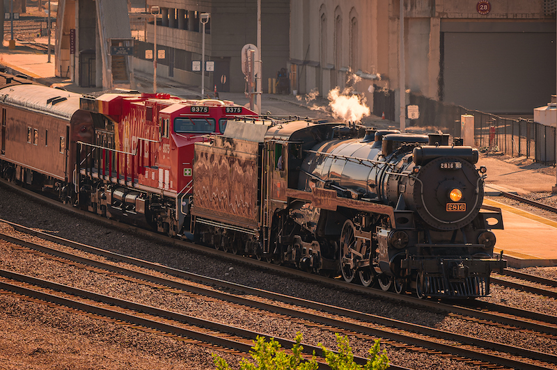 A steam train on track near buildings and trees