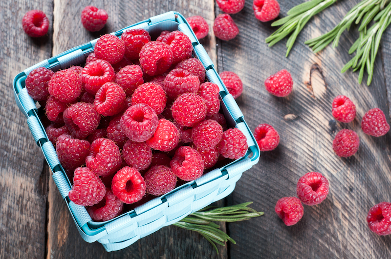 Fresh raspberries in a basket and rosemary on a wooden background.