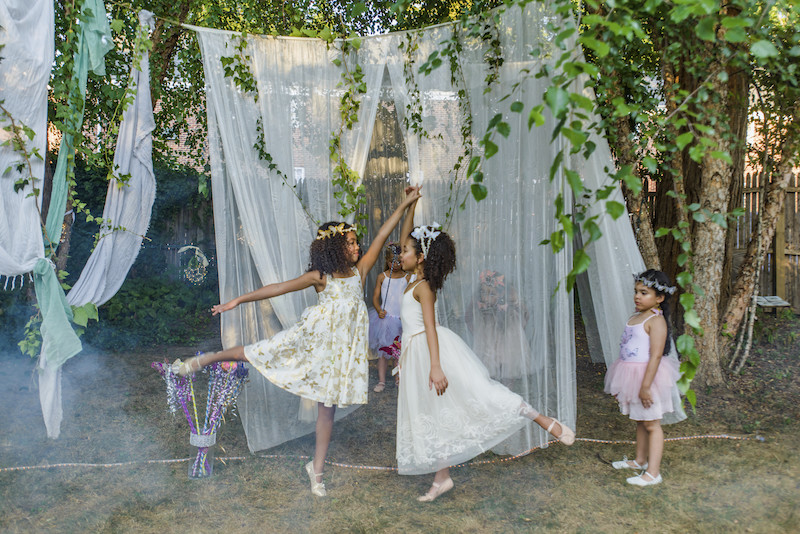 Two young girls, dressed as fairies, dancing outdoors, younger girl watching from side