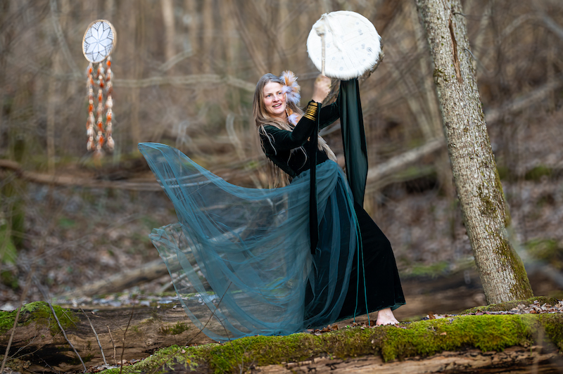 Woman in green dress playing a drum while laying on a fallen log in a forest.