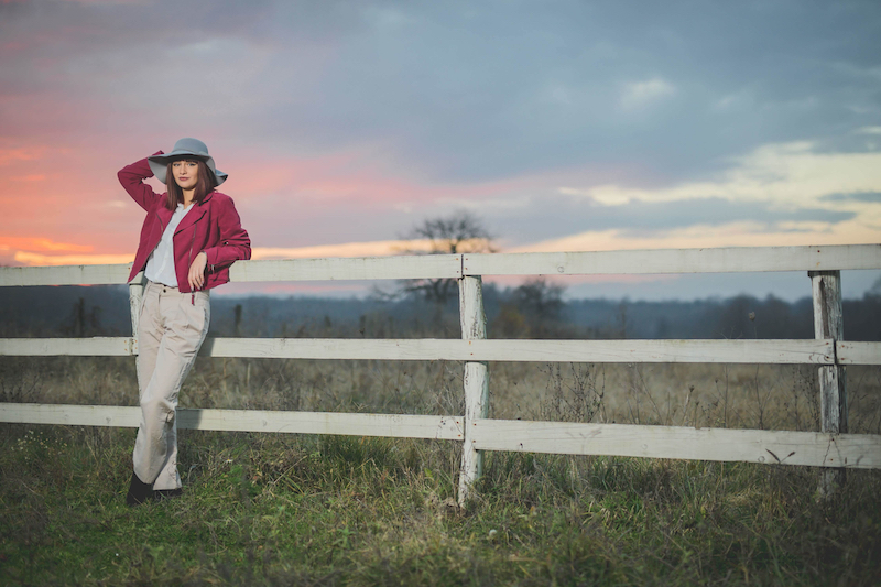 A Caucasian female from Bosnia Herzegovina wearing a purple jacket posing near wooden fence at sunset