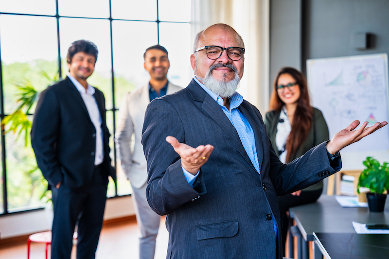 Closeup portrait of senior Indian Asian business professional or CEO standing confidently with folded hands and a smile, colleagues blurred in the background