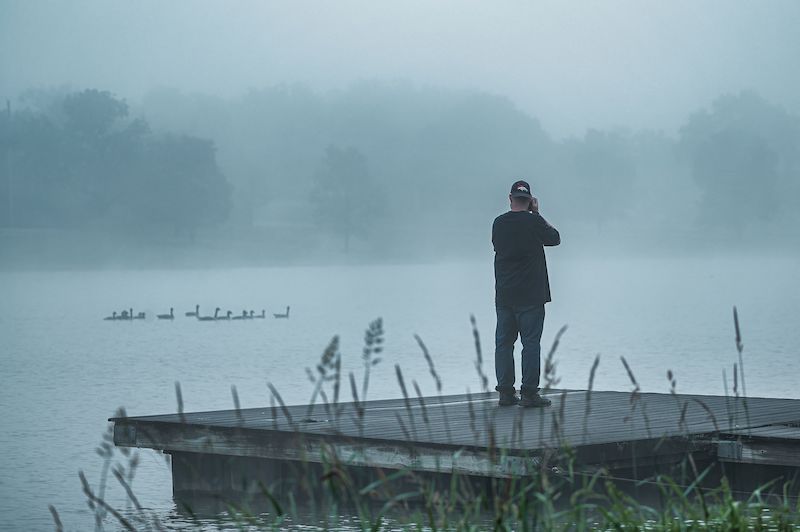 A man standing on a dock by a foggy lake in the early morning