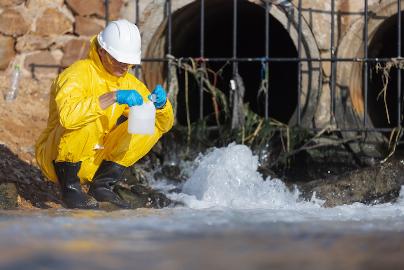 A scientist in yellow protective gear collects a water sample in a plastic bottle near a drainage pipe. The scene highlights environmental monitoring and pollution testing at an industrial outfall.