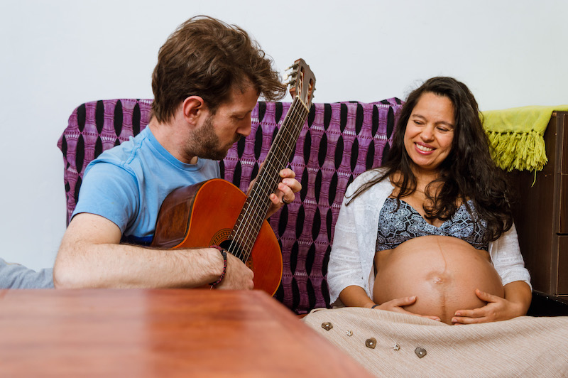 man playing the acoustic guitar for his baby that is inside the womb of his wife, sitting on the sofa inside the house, enjoying together.
