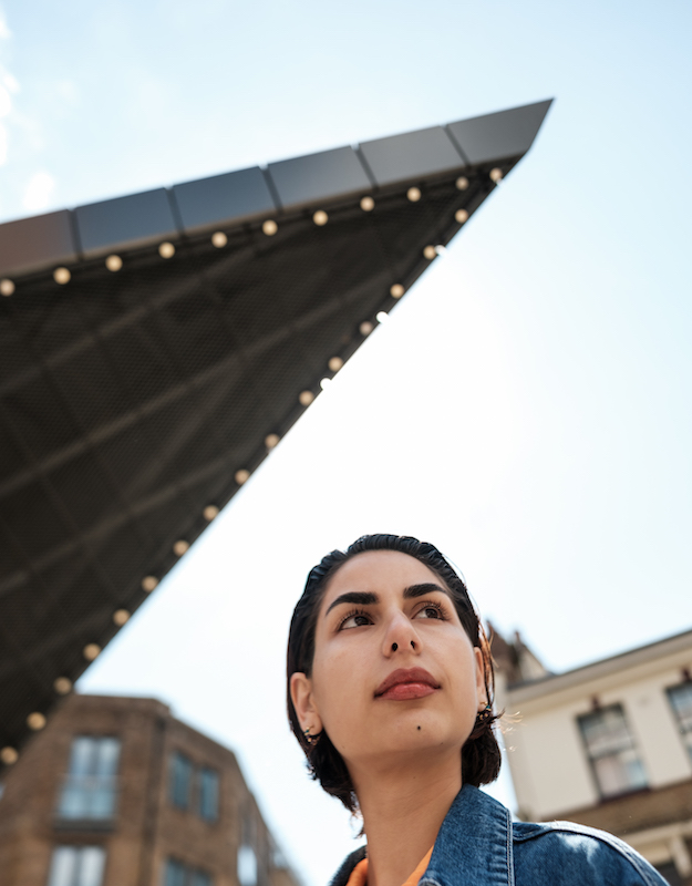 Stylish young woman walking through a modern city, gazing up at a striking triangular architectural feature against a clear blue sky, appreciating urban design