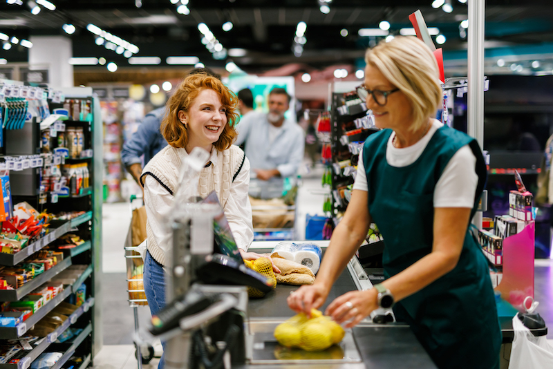 Cheerful young woman buying groceries at a supermarket checkout, talking with the cashier who is scanning her purchases on a conveyor belt