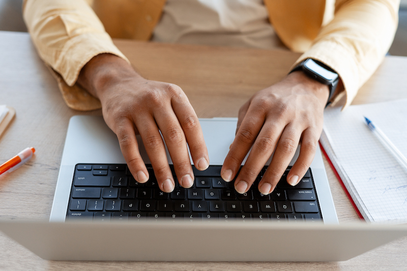 Person's hands typing on a laptop keyboard at a wooden desk, working, studying, or communicating online